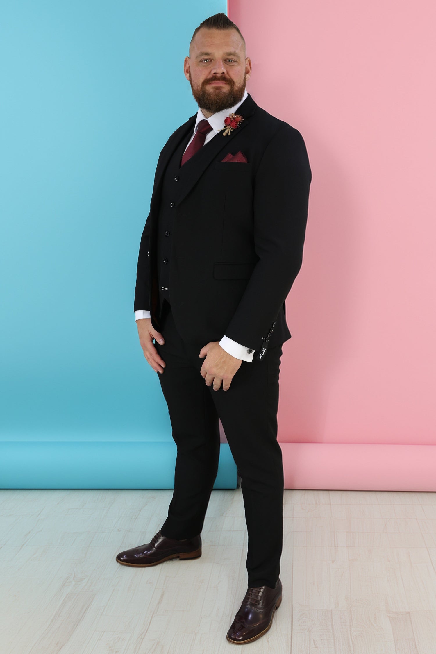 Man wearing a tailored black three-piece suit with a maroon tie and matching pocket square, complemented by gold buttons and brown dress shoes. The suit offers a sharp, sophisticated look suitable for formal events