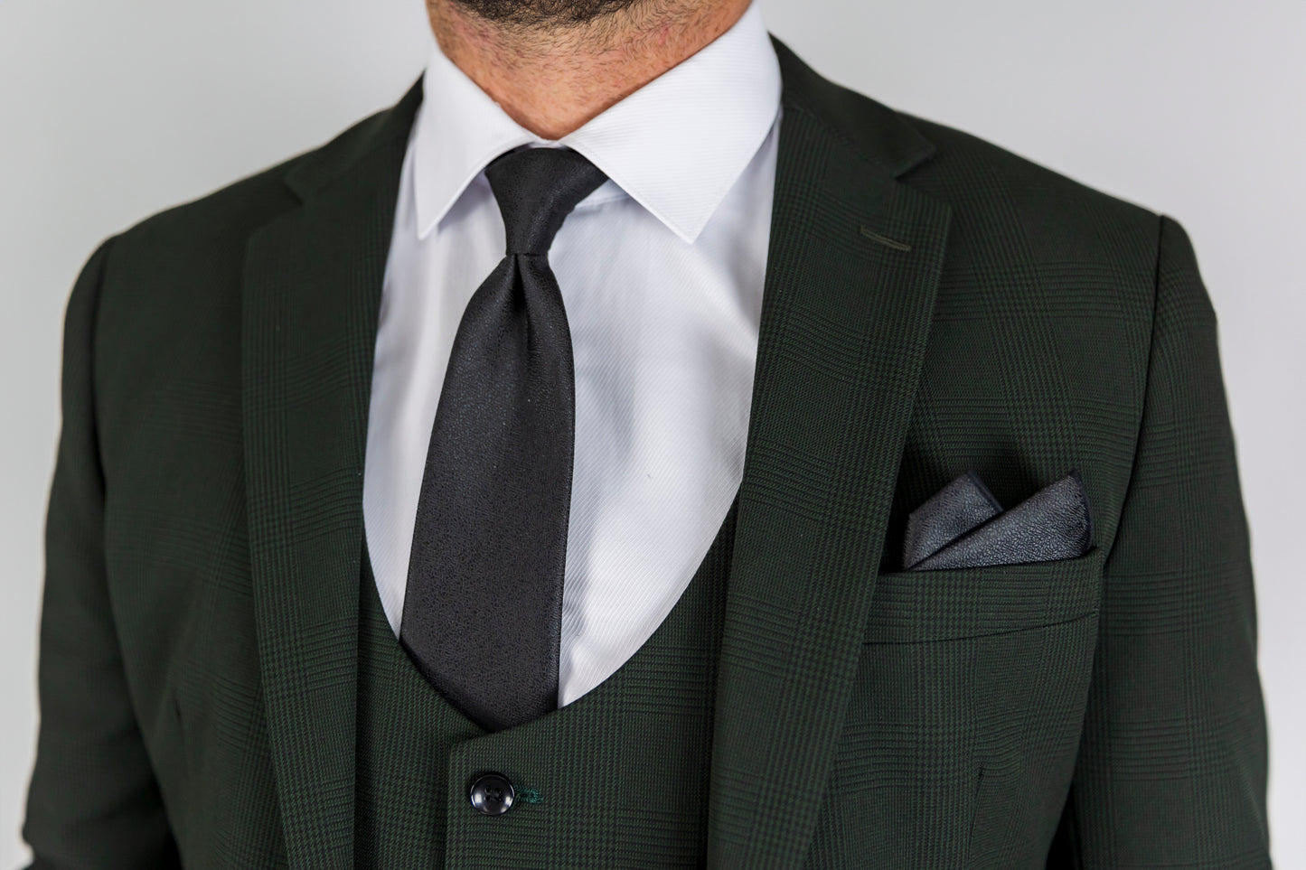 A close-up view of the Suitbae Charles green suit, showcasing the elegant texture and detail of the fabric. The suit is paired with a crisp white shirt and a sleek black tie, creating a sophisticated contrast. A matching dark pocket square is neatly folded in the breast pocket, complementing the overall look. The subtle pattern of the suit adds depth, while the deep green colour offers a modern twist to a classic design.