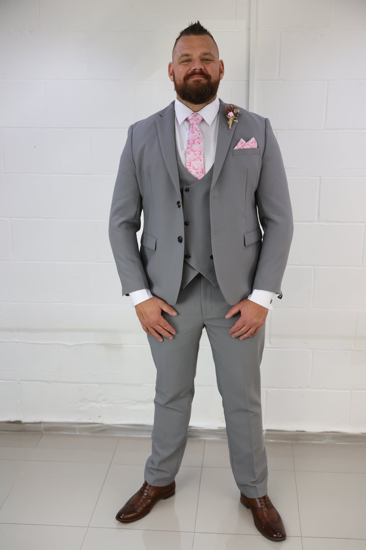 Model wearing the SUITBAE Mason Grey Three Piece Suit with pink floral accessories — a sharp, fitted wedding suit ideal for grooms, groomsmen, or summer formal events.