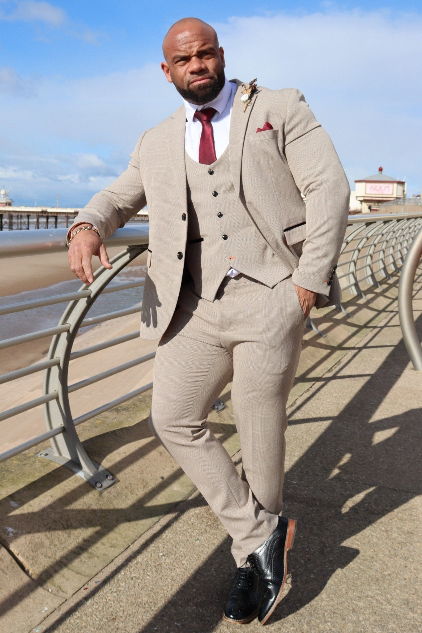 Man standing on Blackpool promenade wearing the SUITBAE Charles Beige Three-Piece Suit. The beige checked suit features contrast black piping, a horseshoe-cut waistcoat, and tangerine lining — paired with a burgundy tie and pocket square for a smart, modern groom look.