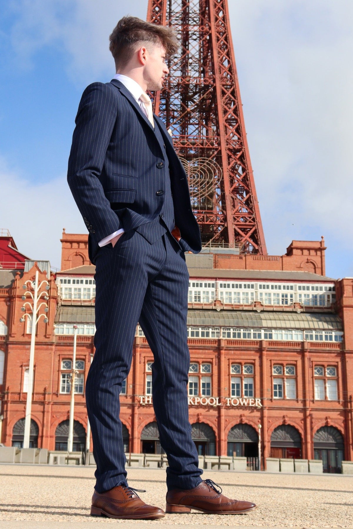 Man wearing a navy pinstripe three-piece suit with a white shirt, cream tie, and brown brogues, standing confidently in front of the Blackpool Tower. The suit’s tailored fit and fine pinstripe detailing stand out against the red-brick landmark backdrop.