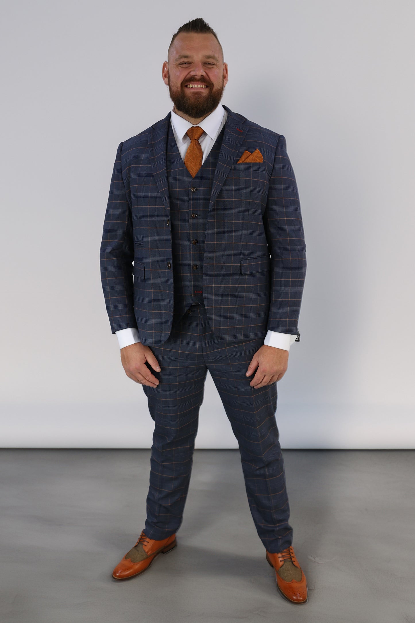 Model wearing the SUITBAE Thomas Check Three Piece Suit in navy with burnt orange overcheck, styled with matching orange tie and pocket square — perfect for weddings, races, or formal events.