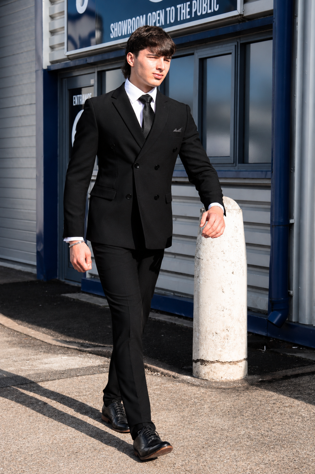 A man wearing a black double-breasted suit, white dress shirt, and black tie walks outdoors in front of a building with blue trim and a concrete bollard.