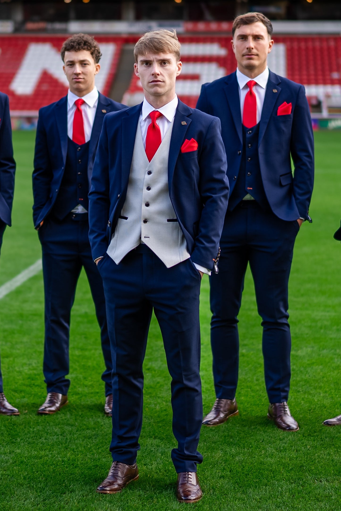 Barnsley FC captain wearing the SUITBAE Charles Navy Contrast Three-Piece Suit with a light grey waistcoat, red tie, and matching pocket square, styled with brown brogues. He stands confidently on the pitch with teammates in matching navy suits behind him.