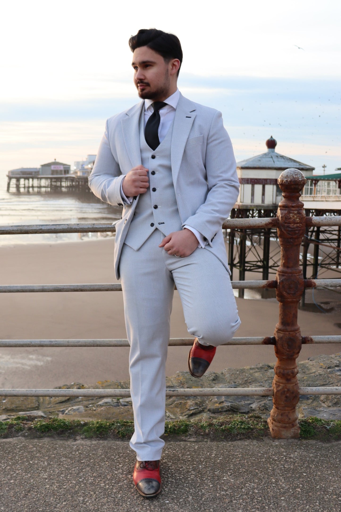 Model wearing the SUITBAE Charles Stone Three-Piece Suit in light stone grey with a black tie, posing by Blackpool pier — ideal for weddings, races, and prom season.