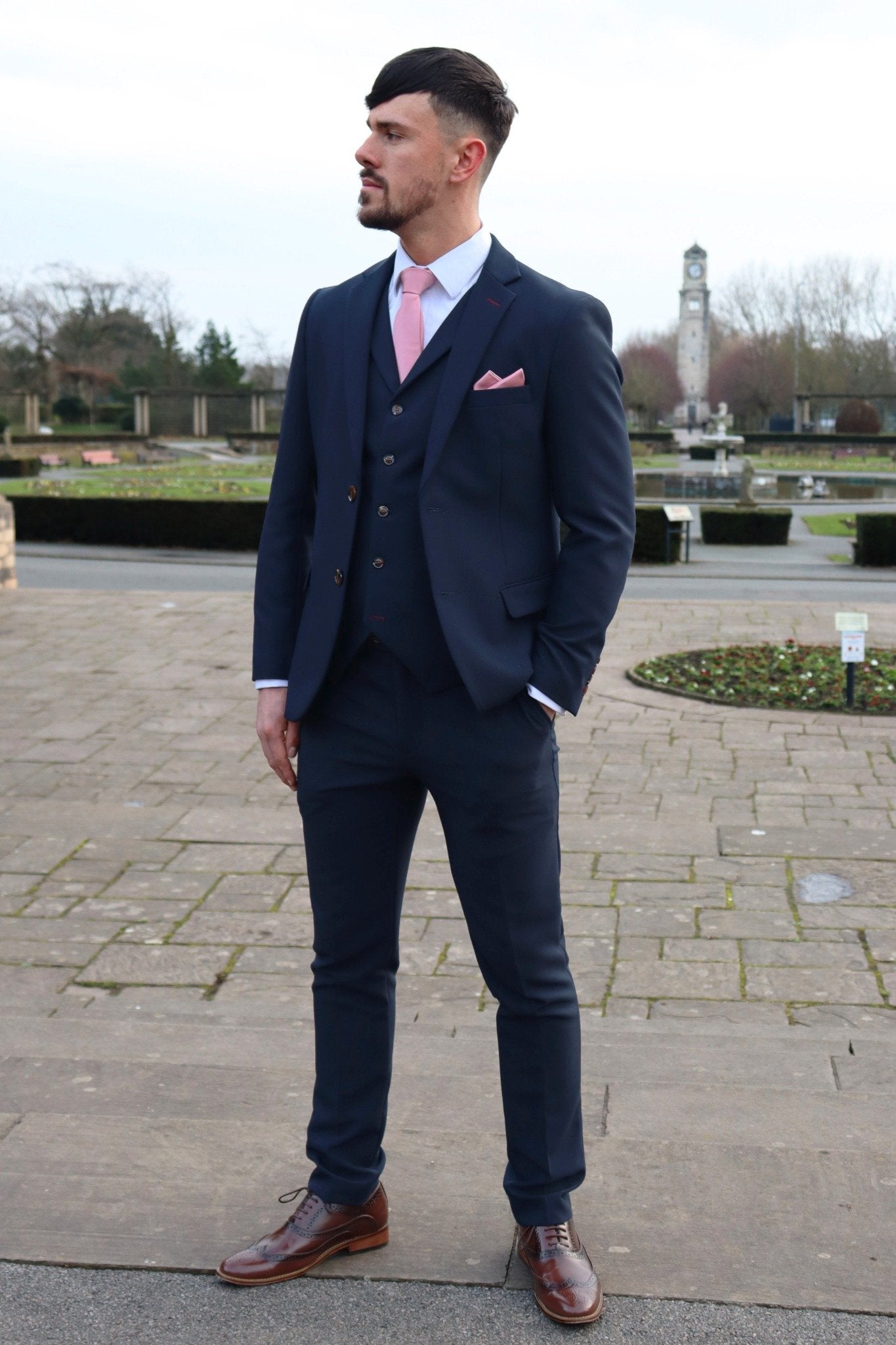 Man standing outdoors wearing a navy three-piece suit with a pink tie and pocket square, paired with brown brogues, posing confidently in a park with a clock tower in the background — part of the SUITBAE formalwear collection.