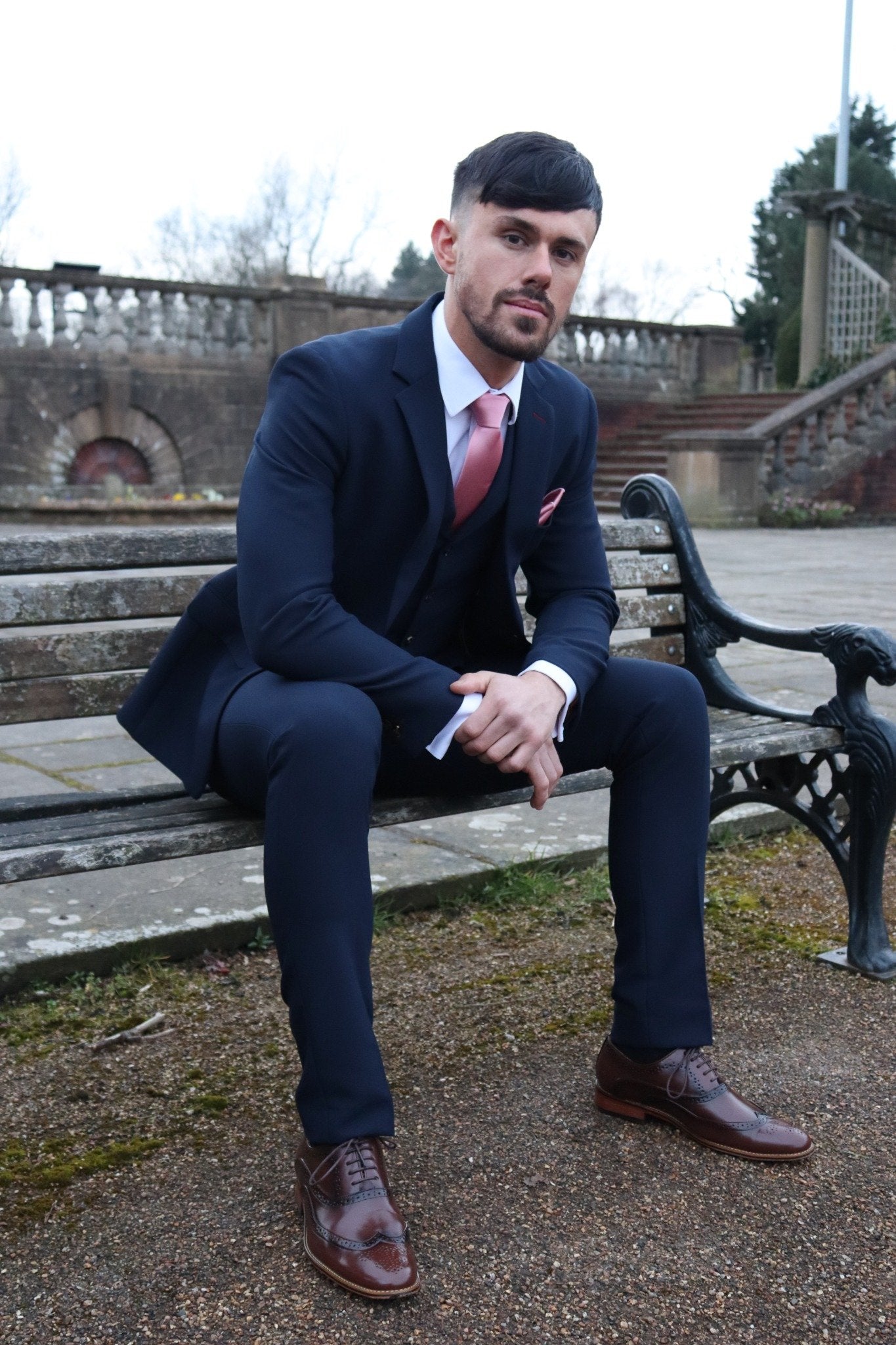 Man sitting on a park bench wearing a navy three-piece suit with a pink tie and pocket square, paired with brown brogue shoes — styled as part of the SUITBAE formalwear collection.