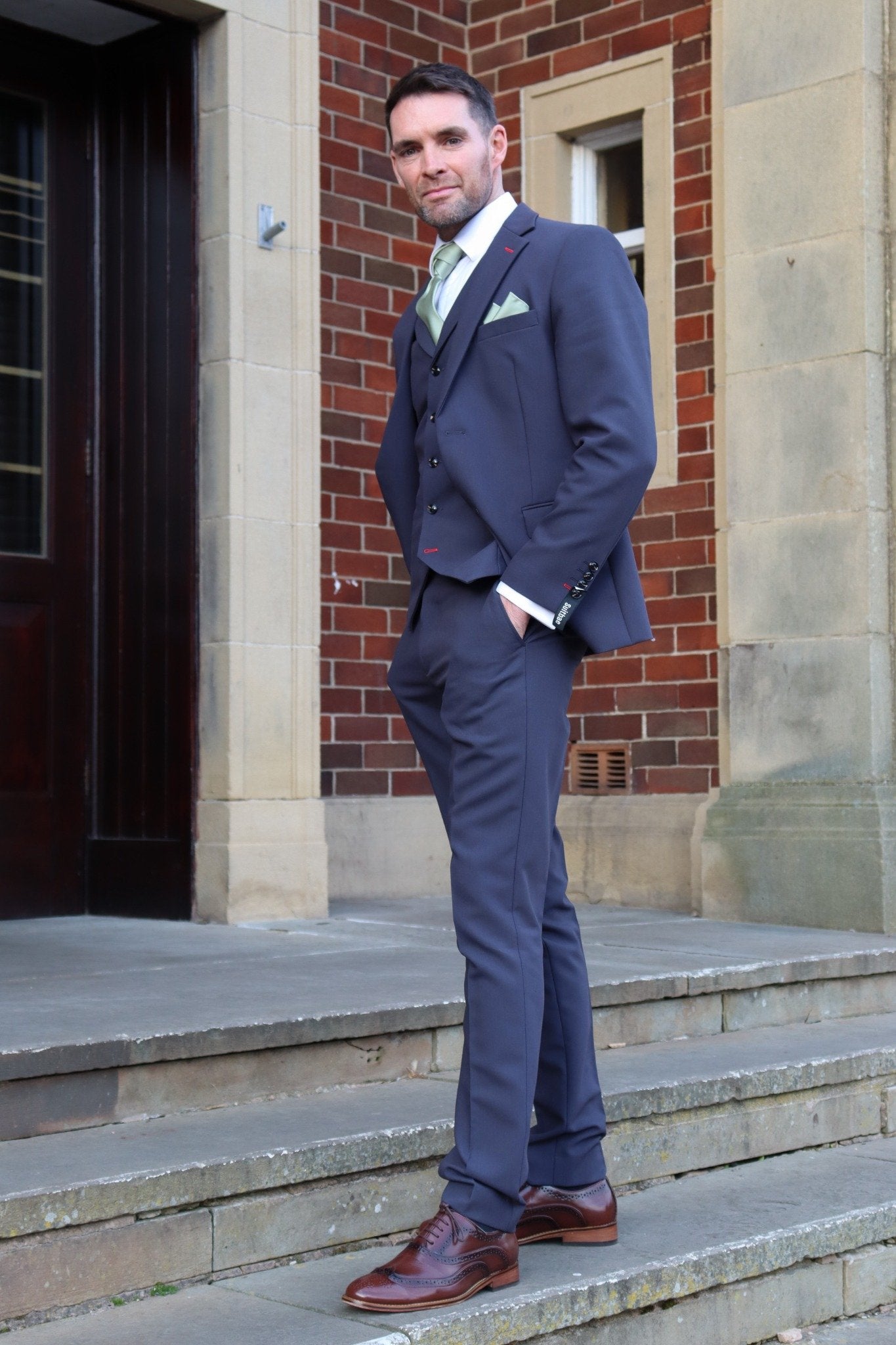 Man confidently standing on stone steps outside a brick building wearing a midnight blue three-piece suit with a sage green tie and pocket square, styled with brown brogue shoes — from the SUITBAE formalwear collection.