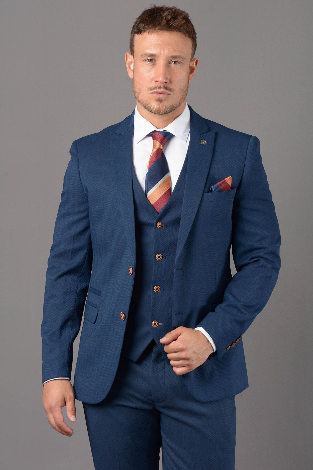 Man wearing a tailored royal blue three-piece suit with a striped tie and matching pocket square. The suit features brown buttons and offers a modern, sophisticated look perfect for weddings, business, and formal events.