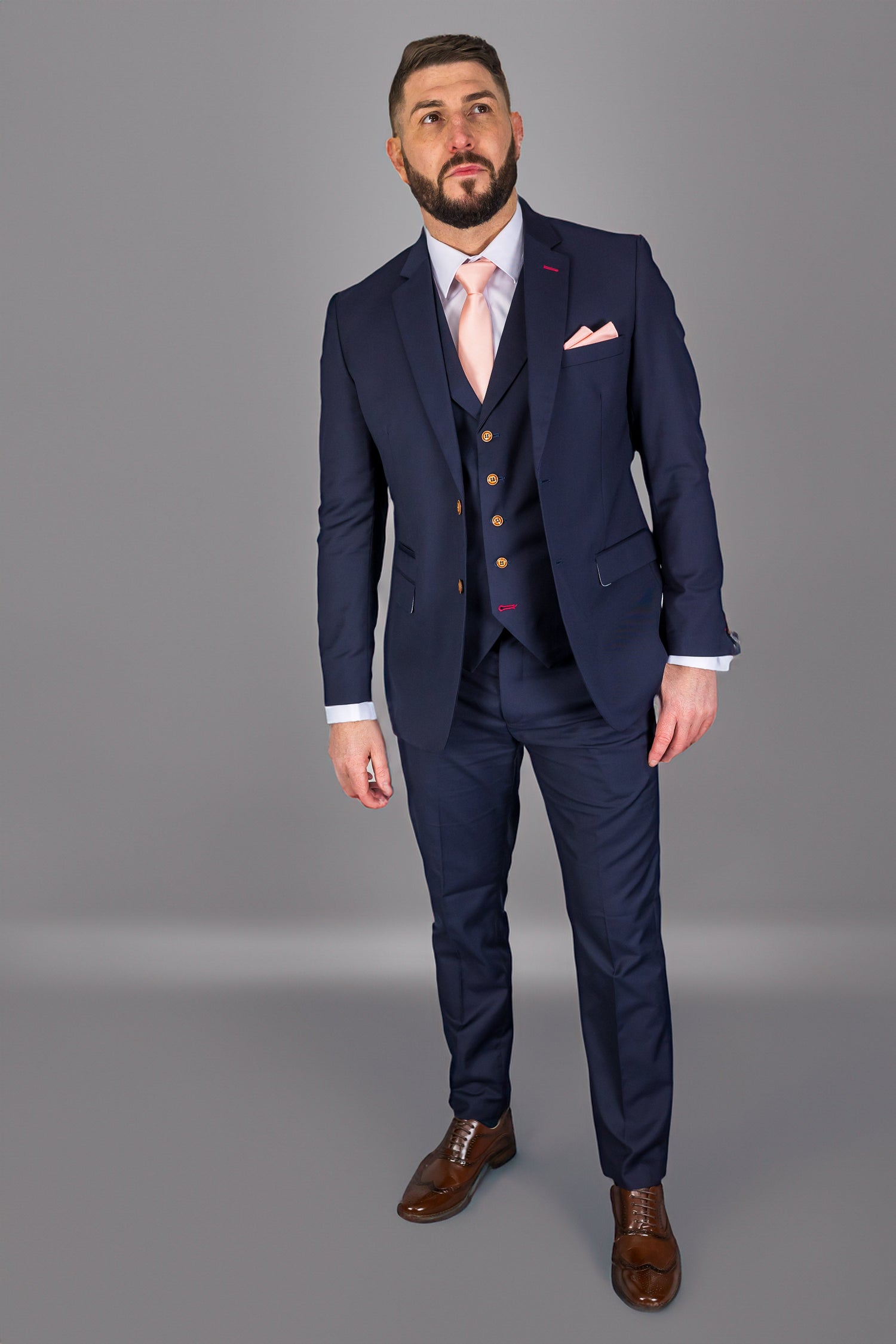 "Man wearing a tailored navy blue three-piece suit with a light pink tie and matching pocket square. The suit features gold buttons and brown dress shoes, creating a modern, stylish look perfect for weddings, business, and formal events.