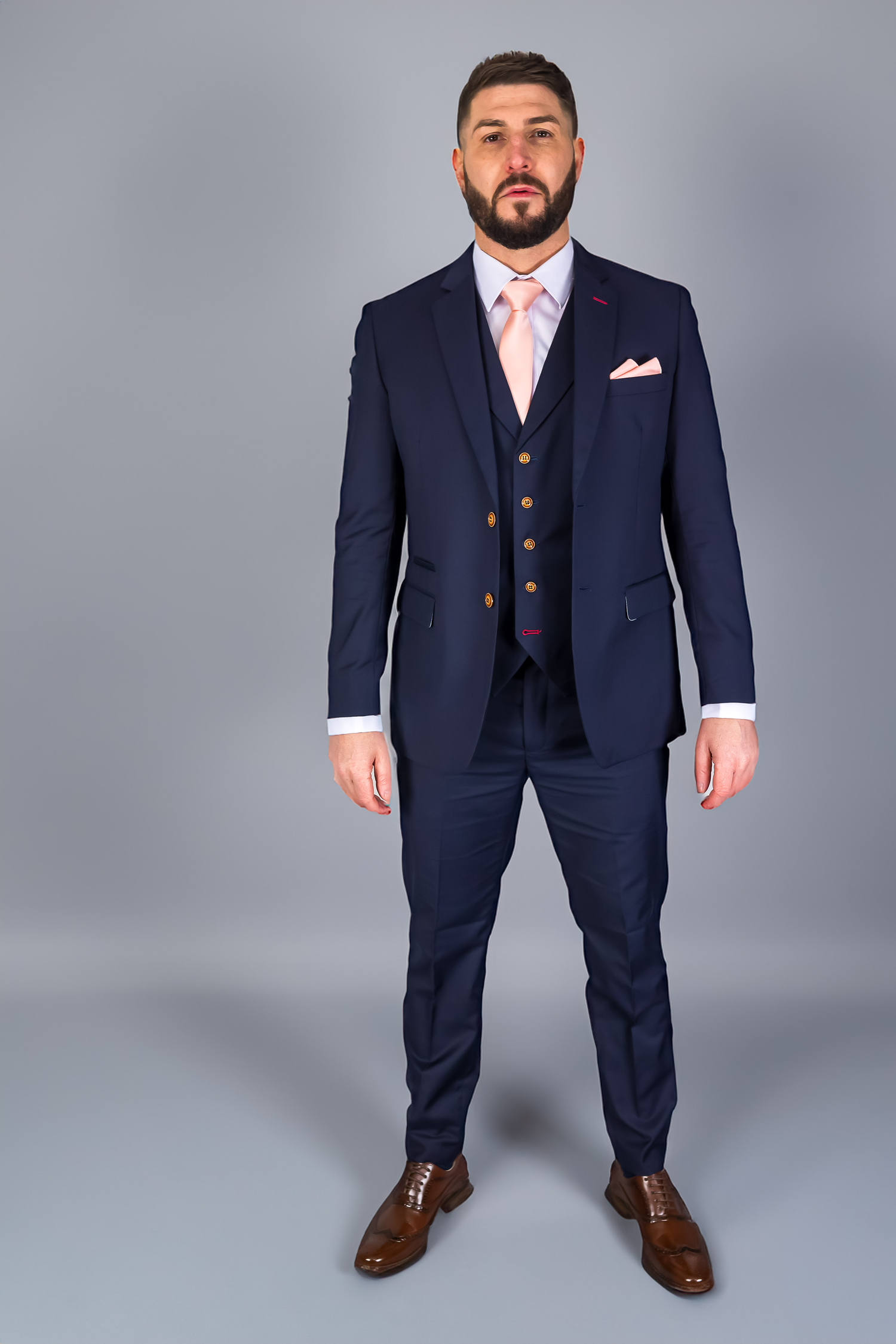 Man wearing a tailored navy blue three-piece suit with a pink tie and matching pocket square, perfect for weddings or formal events. The suit features gold buttons, and he completes the look with brown dress shoes. Modern style for any formal occasion.