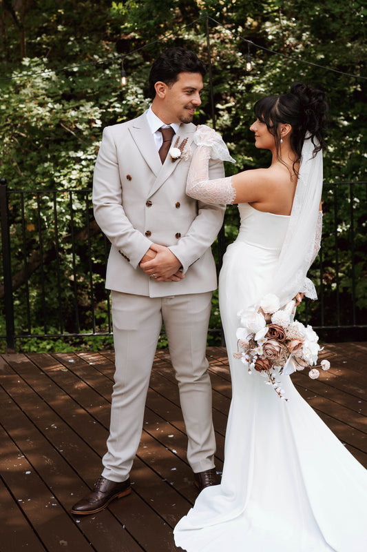 A bride in a strapless white wedding gown with long sheer sleeves and a veil holds a bouquet while standing on a wooden deck with a groom in a light beige double-breasted suit and brown tie, set against a backdrop of green trees.