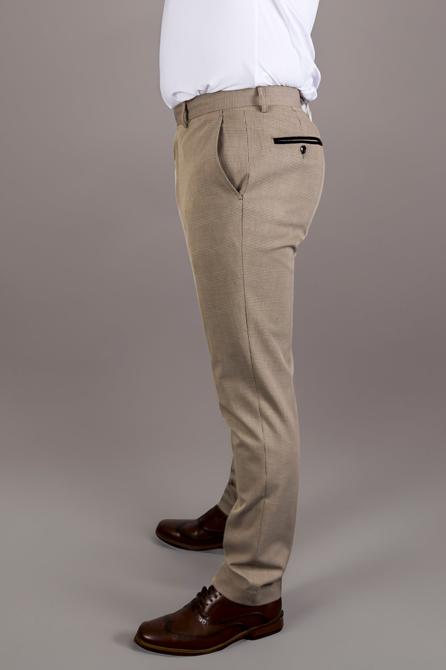 A side view of the Suitbae Charles beige suit trousers, featuring a Prince of Wales check pattern. The slim fit design has front and back pockets with contrasting black piping on the back, enhancing the tailored look. Paired with brown brogue shoes, the trousers provide a refined silhouette, ideal for formal wear, business meetings, or special occasions. The beige colour and subtle pattern add sophistication to the classic menswear style.