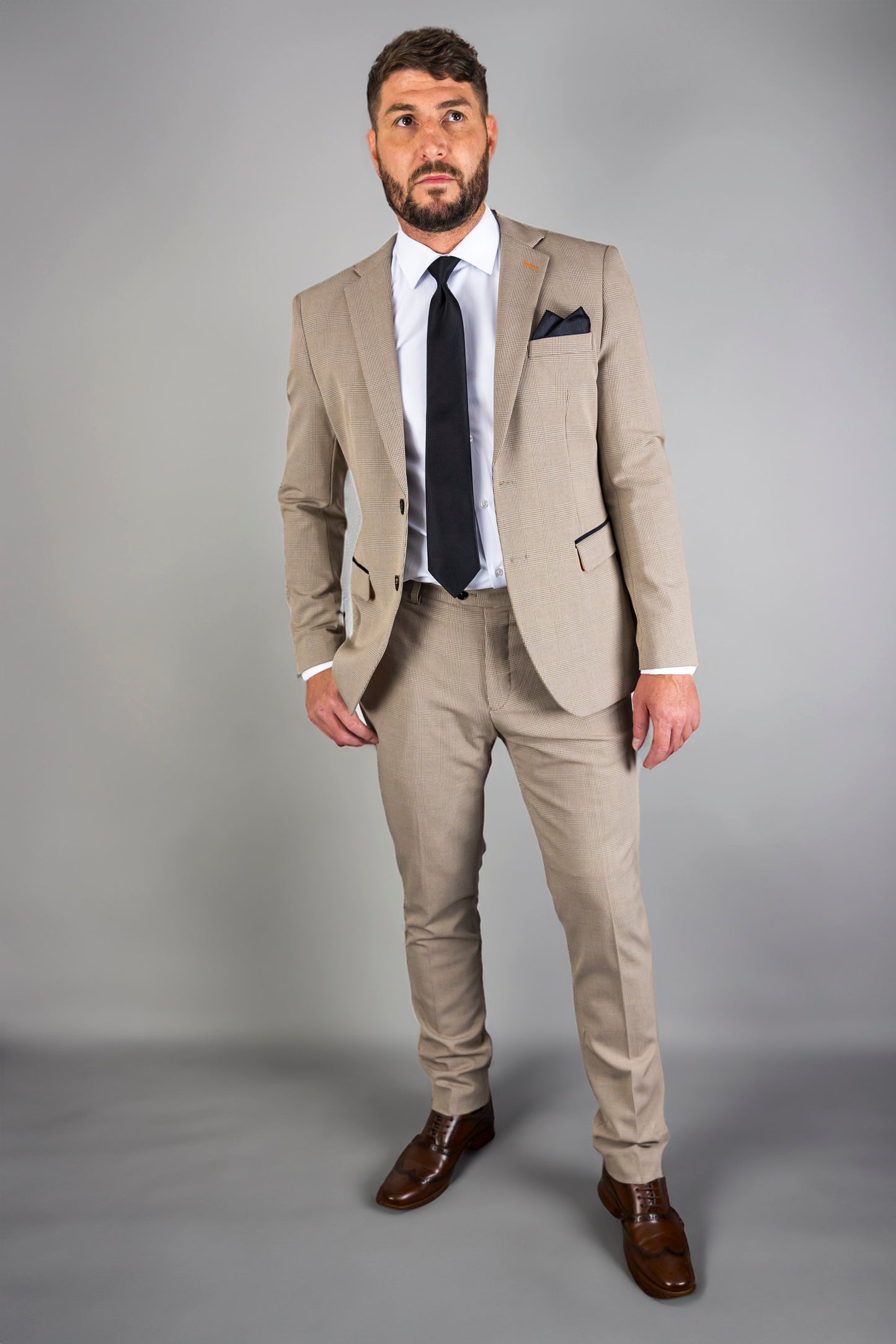 A man models the Suitbae Charles beige suit, featuring a Prince of Wales check pattern. This slim fit, single-breasted suit is paired with a white shirt and a classic black tie for a sophisticated look. The tailored jacket includes notch lapels and a dark pocket square, while matching trousers create a streamlined silhouette. Styled with polished brown brogue shoes, this formal ensemble is perfect for weddings, business meetings, or special events. The modern design and sharp details offer a refined, elegan