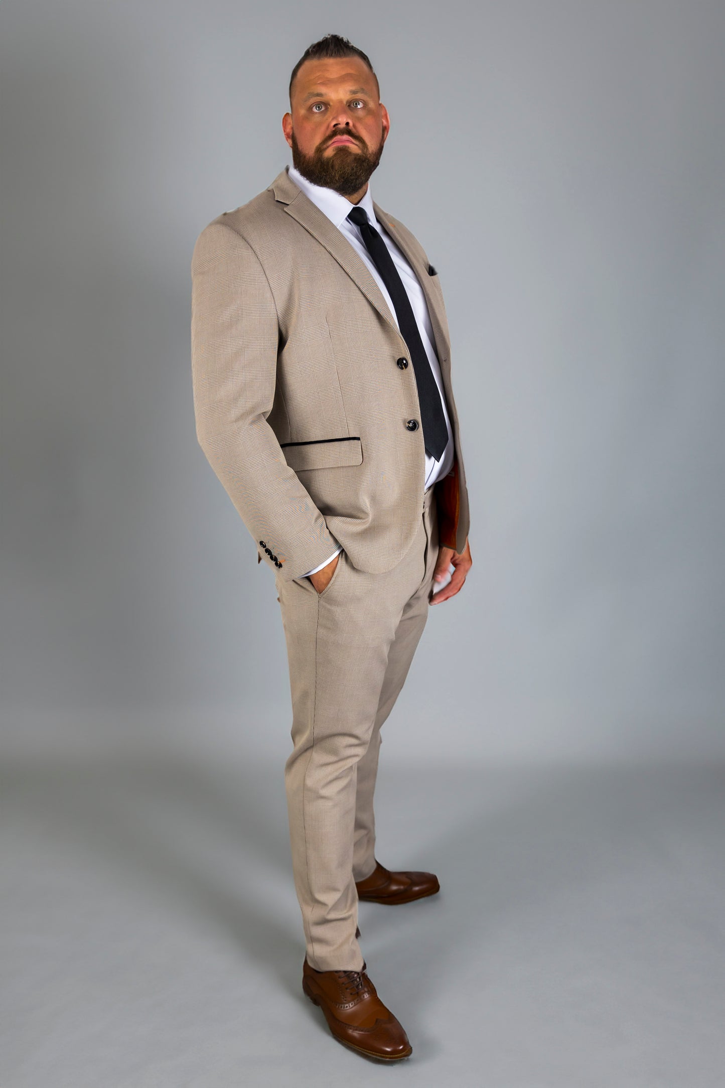 A man models the Suitbae Charles beige suit in a big and tall plus size fit, featuring a Prince of Wales check pattern. The tailored jacket and slim fit trousers create a refined look, paired with a white shirt, black tie, and brown shoes. Designed for a polished appearance for larger frames.