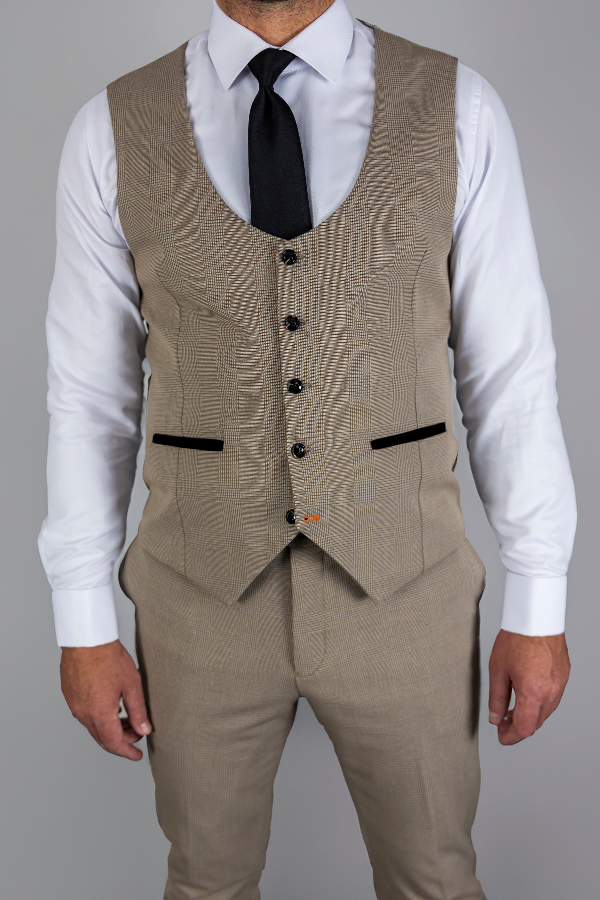 A man wears the waistcoat from the Suitbae Charles beige suit, featuring a Prince of Wales check pattern. This slim fit waistcoat is styled with a white dress shirt and a black tie, showcasing a tailored cut. The design includes contrasting black pockets and detailed buttons, adding a touch of elegance to the classic formalwear. Ideal for weddings, business, or formal events, this waistcoat provides a refined and sophisticated look.