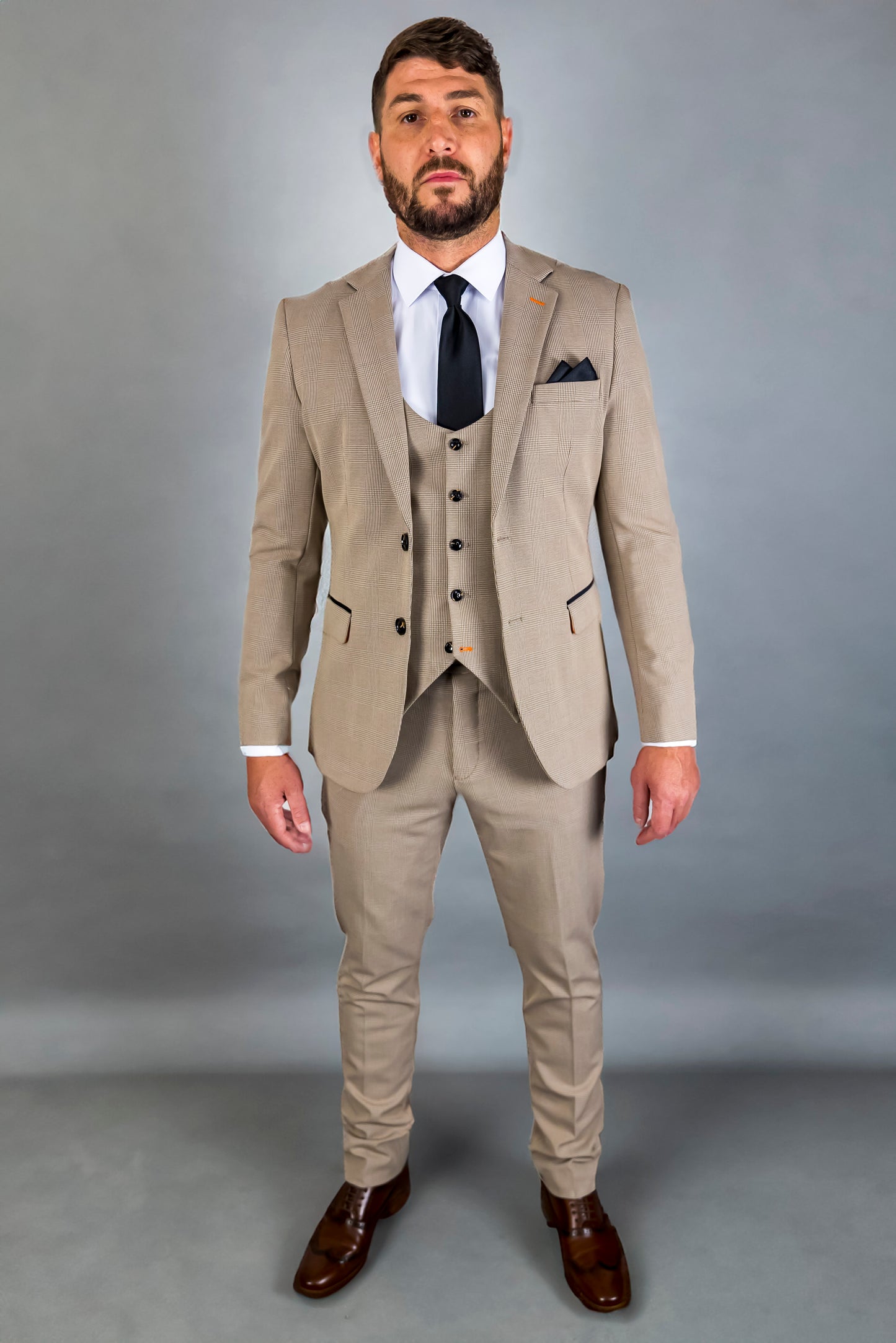 A man models the Suitbae Charles beige suit, featuring a Prince of Wales check pattern. The three-piece slim fit suit includes a tailored jacket, waistcoat, and matching trousers. Styled with a white shirt and a black tie, this ensemble is complemented by a dark pocket square and brown dress shoes. The structured cut and intricate pattern make it ideal for weddings, business attire, or formal occasions, offering a refined yet modern appearance.