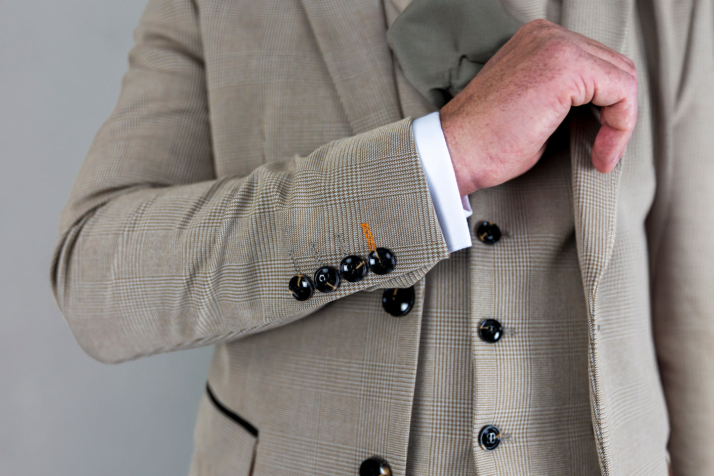 A close-up of the Suitbae Charles beige suit, highlighting the Prince of Wales check pattern and fine tailoring. The jacket sleeve features four black cuff buttons and distinctive contrast stitching, showcasing premium craftsmanship. The matching waistcoat is visible, adding a classic touch to the three-piece slim fit suit. Ideal for weddings, business, or formal events, this beige suit combines timeless elegance with modern detailing.