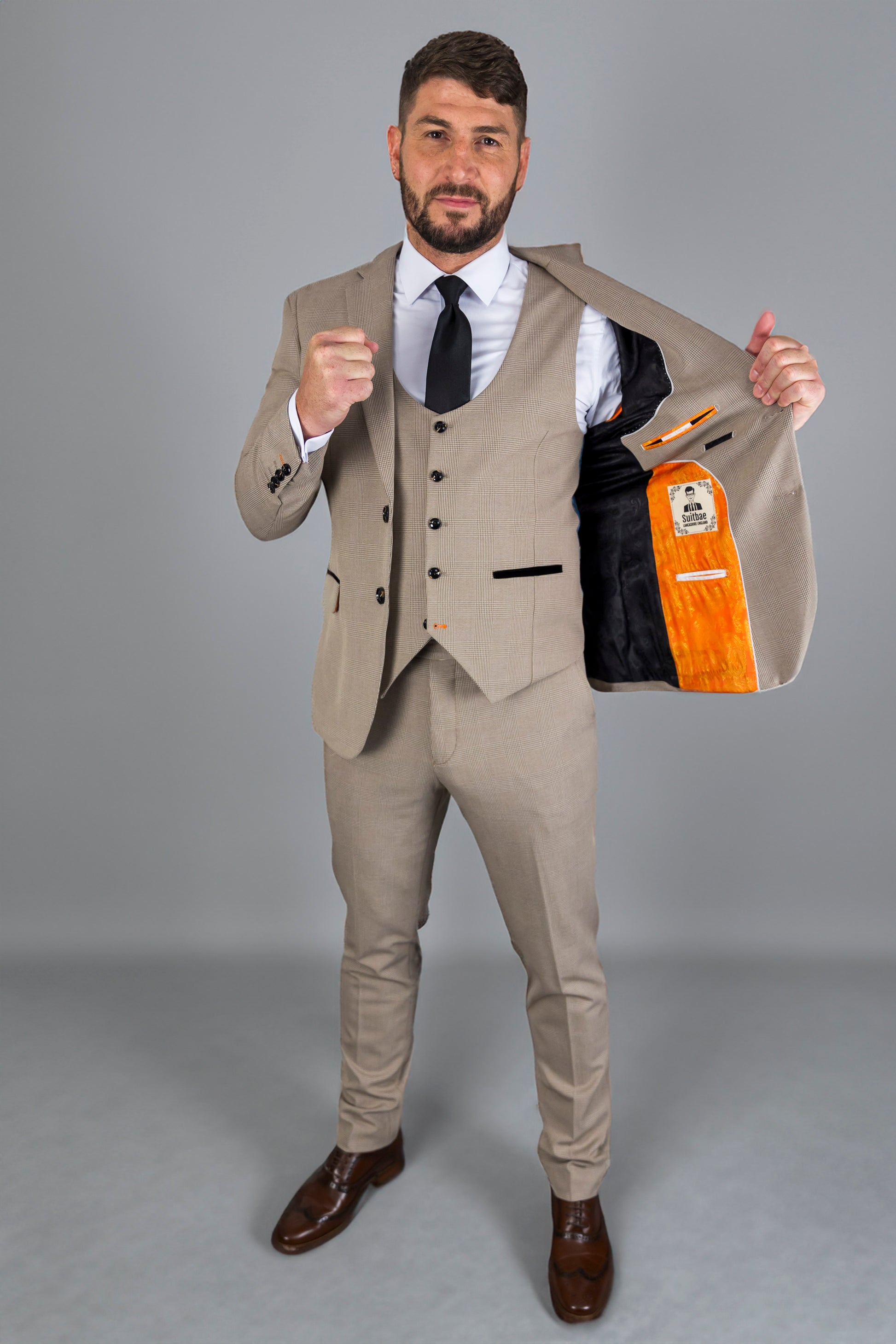 Alt Text: A man models the Suitbae Charles beige suit, featuring a Prince of Wales check pattern. This slim fit, three-piece suit includes a matching waistcoat and trousers, paired with a white shirt and black tie for a classic formal look. The jacket is open, revealing a contrasting orange and black lining with fine stitching details. The suit is styled with polished brown dress shoes, perfect for business wear, weddings, or special occasions. Ideal for a modern, tailored silhouette.