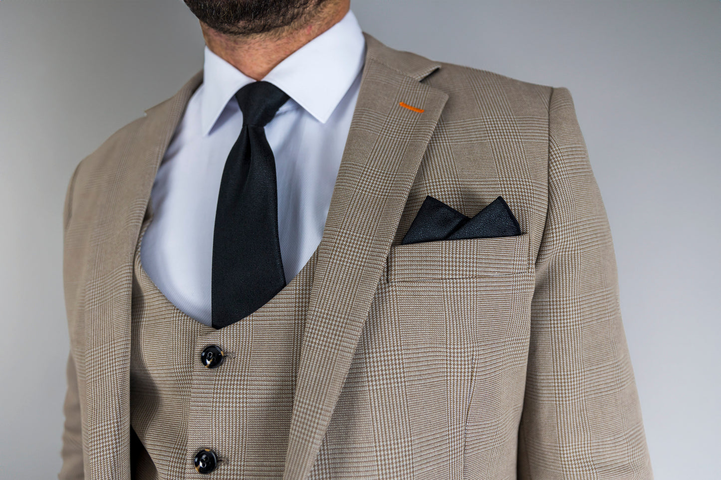 A close-up of the Suitbae Charles beige suit, featuring a Prince of Wales check pattern. The tailored jacket is styled with a white shirt, a black tie, and a dark pocket square for a classic formal look. The waistcoat is visible underneath, showcasing elegant button details. An orange stitch on the lapel adds a subtle accent, highlighting the fine craftsmanship and attention to detail in this modern suit design. Ideal for business, weddings, or special occasions.