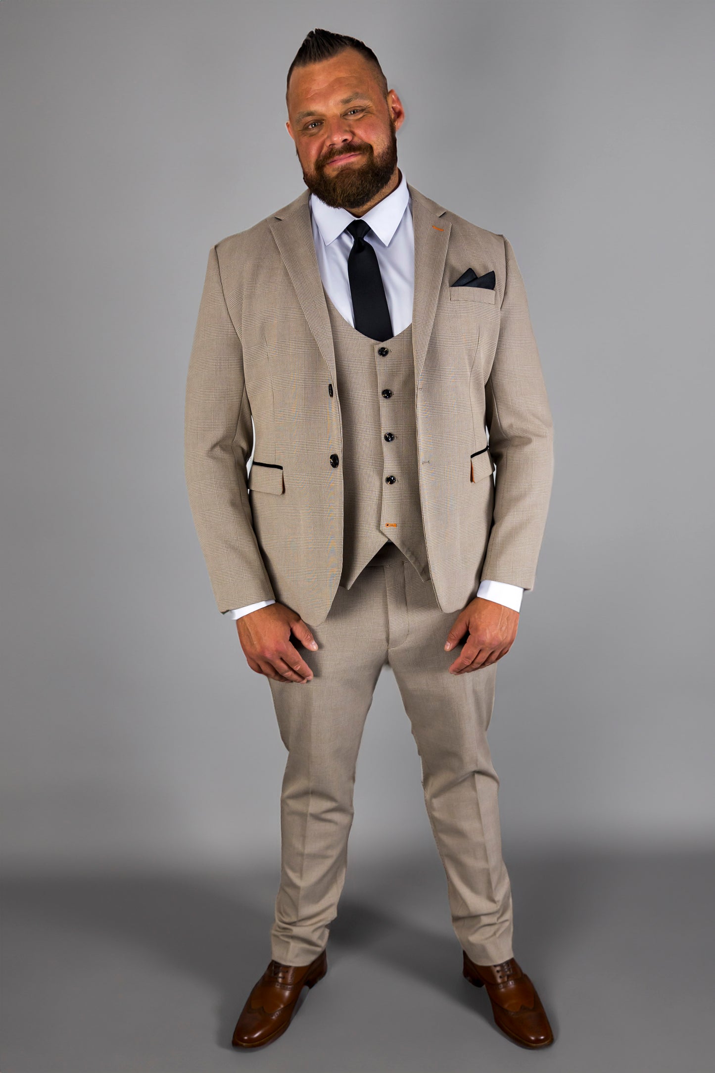 A man models the Suitbae Charles beige suit, featuring a Prince of Wales check pattern. The three-piece slim fit suit includes a matching waistcoat, tailored trousers, and a structured jacket, paired with a white shirt and black tie for a classic formal look. The ensemble is styled with a dark pocket square and polished brown dress shoes, perfect for business meetings, weddings, or other formal events. The beige colour and modern cut create a sophisticated and elegant appearance.