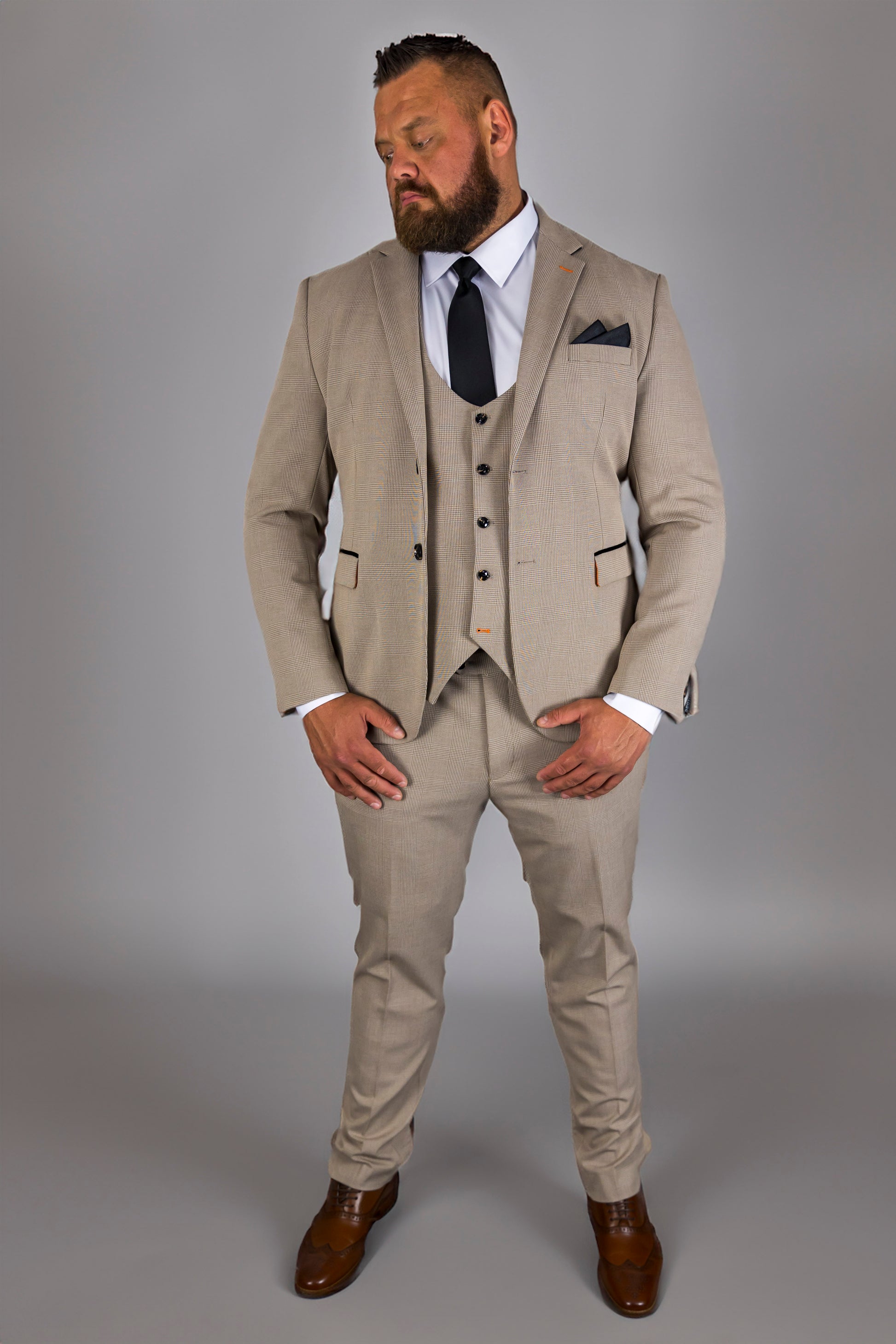 A man models the Suitbae Charles beige suit in a big and tall plus size fit, featuring a Prince of Wales check pattern. This three-piece suit includes a structured jacket, tailored waistcoat, and coordinating trousers, specifically designed to complement larger frames. Paired with a crisp white shirt and black tie, the outfit is finished with a dark pocket square and brown brogue shoes, ensuring a polished look. Perfect for formal occasions, weddings, or business settings, this suit offers a refined style t