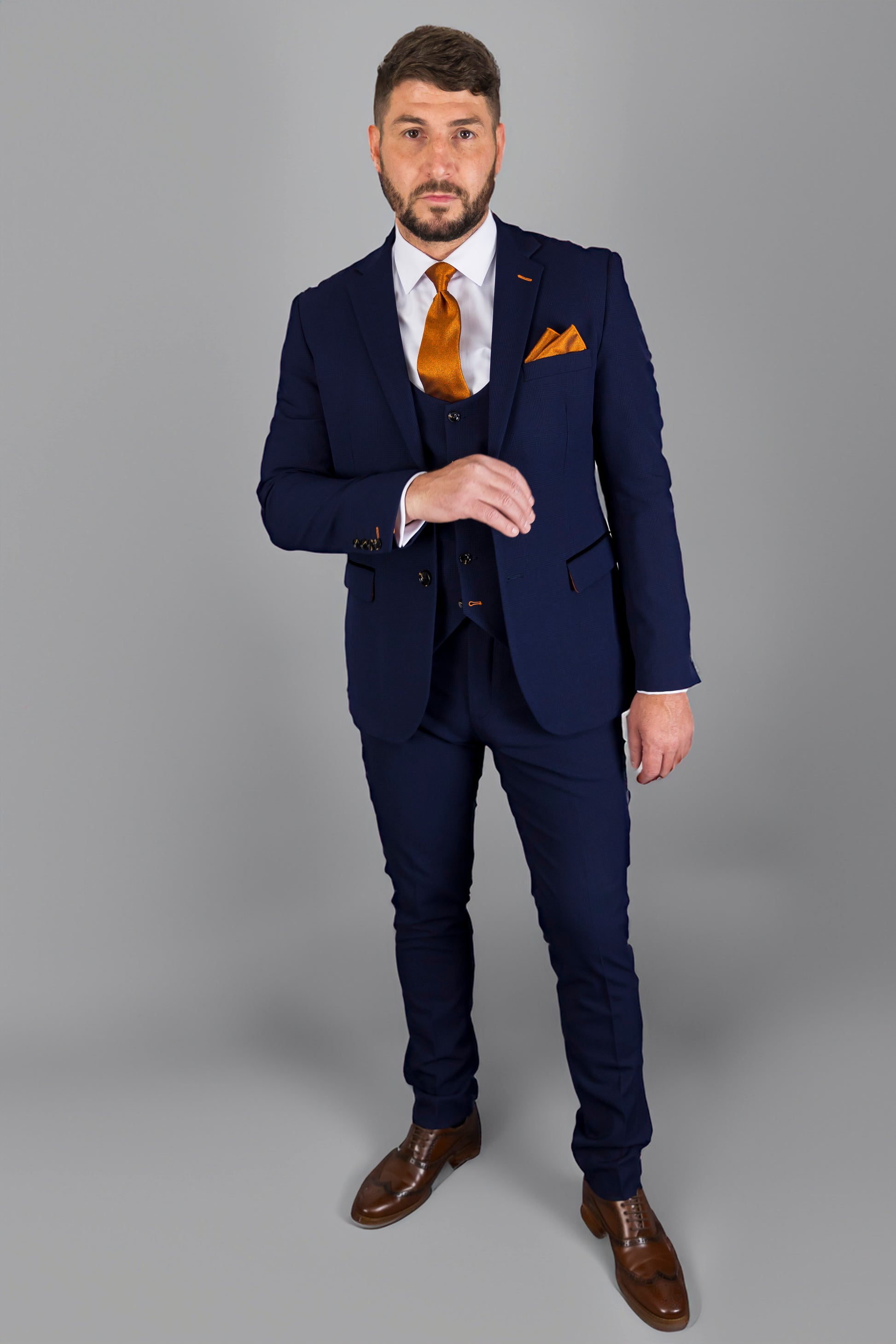 A man is standing in a full-body portrait, dressed in a sharp navy blue three-piece suit. He is wearing a white shirt with an orange tie and a matching orange pocket square. His right hand is slightly raised as if adjusting his cuff. He has short, neatly styled hair and a well-groomed beard. He completes the look with polished brown dress shoes. The background is plain gray, allowing his stylish outfit to stand out.