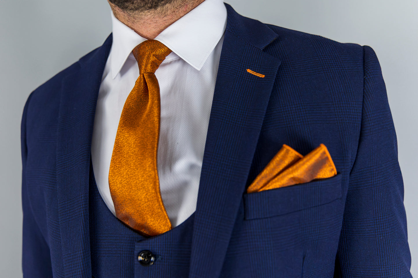 This image shows a stylish, navy check three-piece suit featuring a vibrant orange tie and matching pocket square. The combination of the navy suit and the bold, metallic orange accents creates a striking and modern formal look. The white dress shirt adds contrast, while the subtle check pattern on the suit adds texture. Ideal for formal events, this ensemble balances classic tailoring with contemporary flair, perfect for someone looking to make a statement.