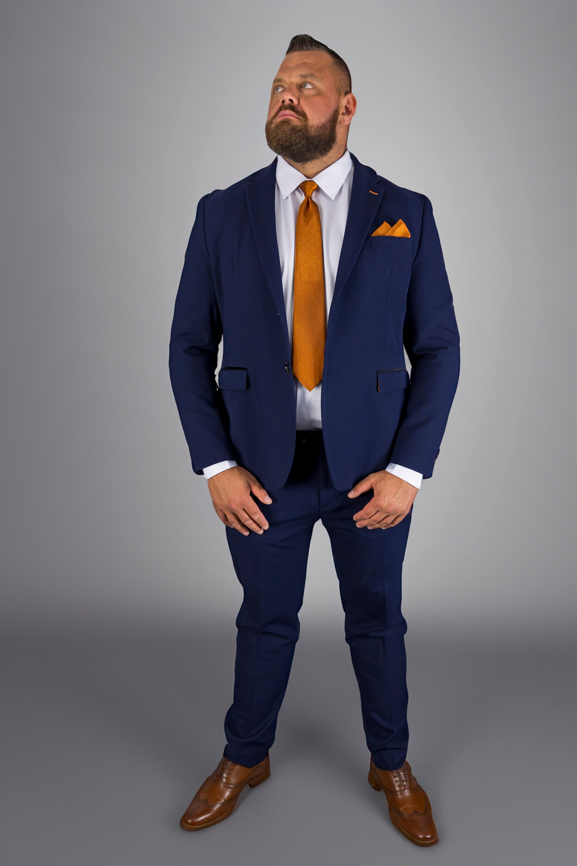 This image shows a man confidently dressed in a navy check three-piece suit, paired with an eye-catching orange tie and pocket square. The well-tailored suit, complete with a crisp white shirt, strikes a perfect balance between formal elegance and modern style. The brown leather shoes complete the sophisticated look, making this outfit ideal for formal occasions, business meetings, or weddings. The overall ensemble demonstrates a powerful sense of fashion with a contemporary twist.