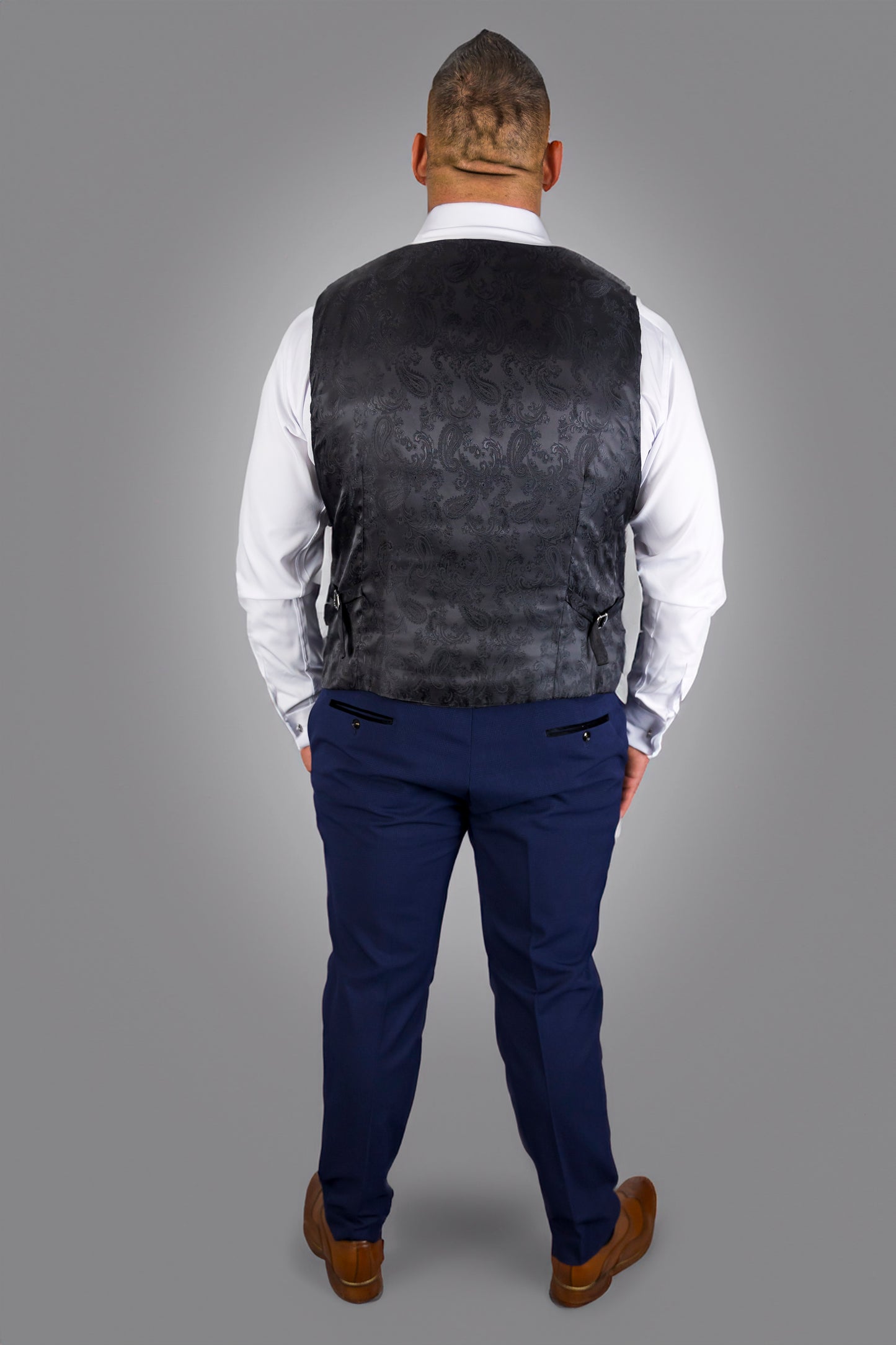 This image shows the back view of a man wearing a navy check three-piece suit with a white dress shirt. The waistcoat features a detailed black damask pattern on the back, adding an extra touch of elegance. The trousers have a tailored fit and are paired with brown leather shoes, completing the formal look. This refined, yet contemporary style makes it ideal for formal events and occasions where a polished appearance is key.