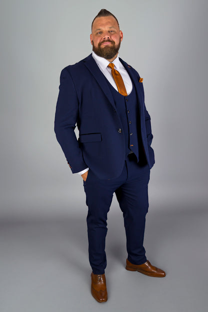 A man is standing confidently in a full-body portrait. He is wearing a tailored navy blue three-piece suit with a white shirt and an orange tie, complemented by a matching pocket square. He has a neatly groomed beard and a mohawk-style haircut. His hands are casually placed in his pockets, and he is wearing polished brown dress shoes. The background is a simple light gray, allowing the sharp contrast of his outfit to stand out.