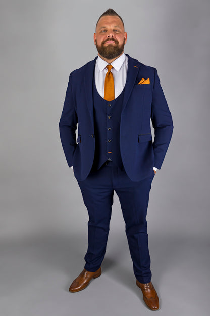 This image shows a confident man wearing a stylish navy Charles check three-piece suit. The suit is complemented by an eye-catching orange silk tie and pocket square, offering a sharp contrast to the deep blue fabric. The suit is tailored for a comfortable fit, with the jacket, waistcoat, and trousers creating a sophisticated, coordinated look. Paired with brown leather shoes, this ensemble is ideal for formal occasions, business meetings, or weddings, blending elegance with modern style.