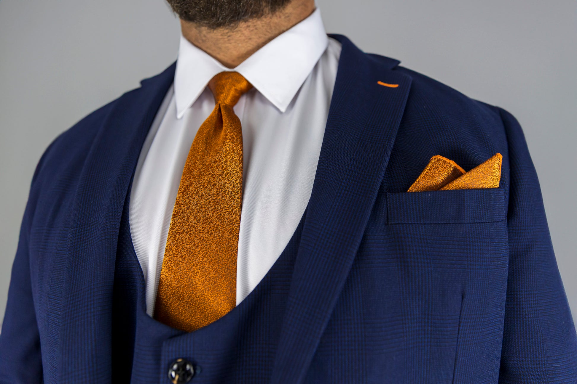 This close-up image highlights a navy check three-piece suit with a striking orange silk tie and matching pocket square. The subtle check pattern on the suit adds texture, while the sharp white dress shirt provides a crisp contrast. The tailored blazer, waistcoat, and trousers complete a polished, formal look perfect for weddings, business meetings, or special events. The bold orange accents give the outfit a modern twist, making it both stylish and sophisticated for the fashion-forward man.