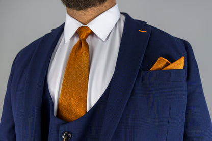 This close-up image highlights a navy check three-piece suit with a striking orange silk tie and matching pocket square. The subtle check pattern on the suit adds texture, while the sharp white dress shirt provides a crisp contrast. The tailored blazer, waistcoat, and trousers complete a polished, formal look perfect for weddings, business meetings, or special events. The bold orange accents give the outfit a modern twist, making it both stylish and sophisticated for the fashion-forward man.