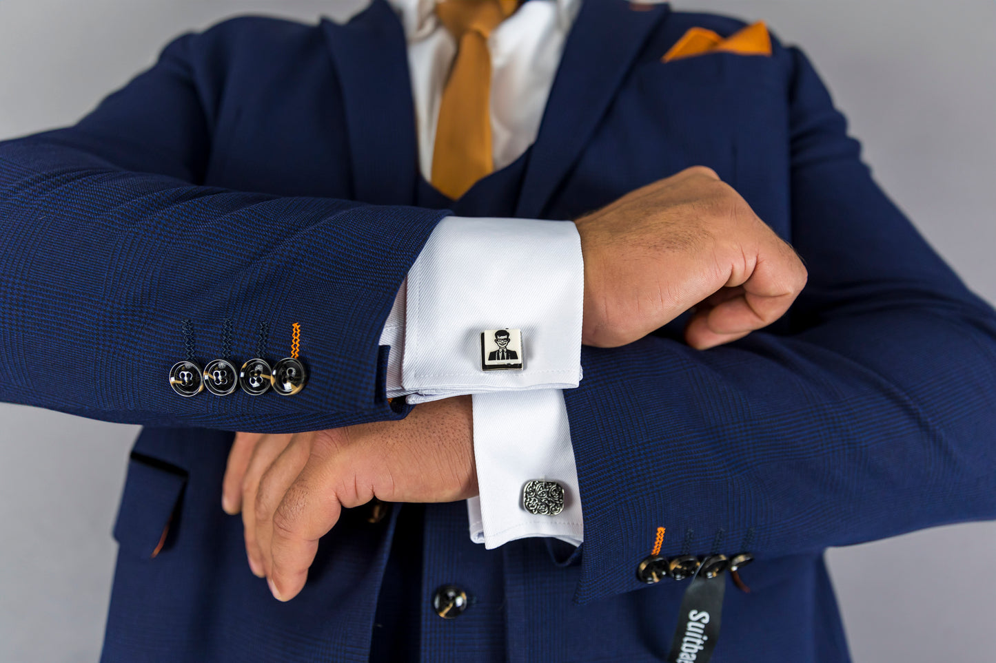 This image showcases the intricate details of a navy check three-piece suit. The close-up focuses on the sleeve, where four buttons are sewn with a distinctive orange thread accent. The man is wearing white dress cuffs adorned with elegant cufflinks, one of which features a detailed portrait. The polished look, complemented by the vibrant orange tie and pocket square, highlights the suit's sharp tailoring and attention to detail, making it perfect for formal occasions or high-profile events.