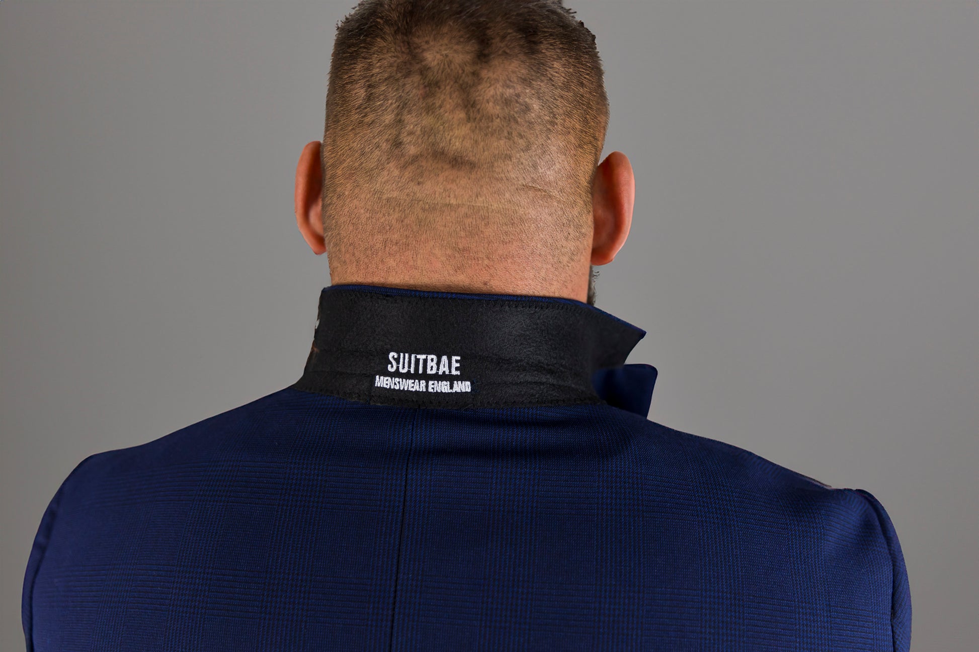 This image shows the back view of a man wearing a navy check suit with the collar turned up, revealing the Suitbae label embroidered in white. The label reads "SUITBAE MENSWEAR ENGLAND" on a black velvet undercollar, adding a sleek, modern touch to the design. The deep blue suit fabric is visible, featuring a subtle check pattern, and this branding detail enhances the premium and tailored feel of the suit.