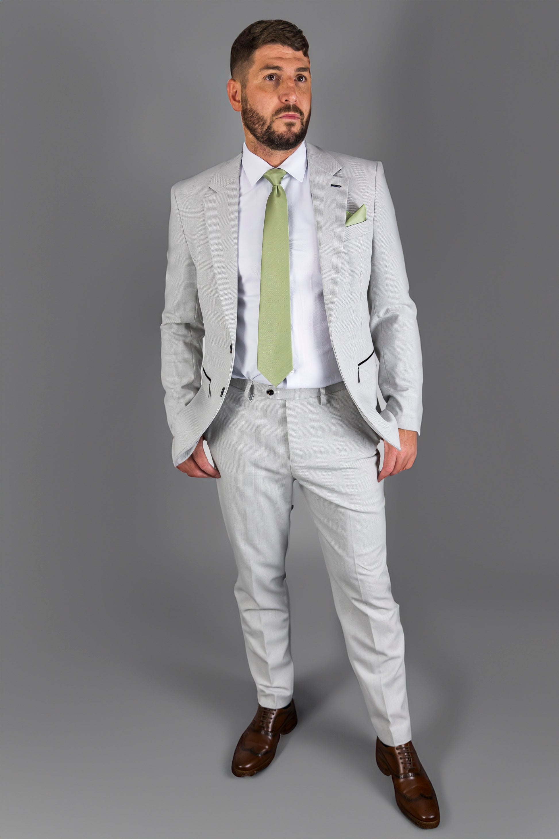 A man wearing the Suitbae Charles stone two-piece suit featuring a classic Prince of Wales check pattern. The outfit includes a tailored jacket and matching trousers paired with a crisp white shirt and a light green tie. Finished with a matching green pocket square and brown formal shoes, creating a sophisticated look perfect for formal events and weddings.