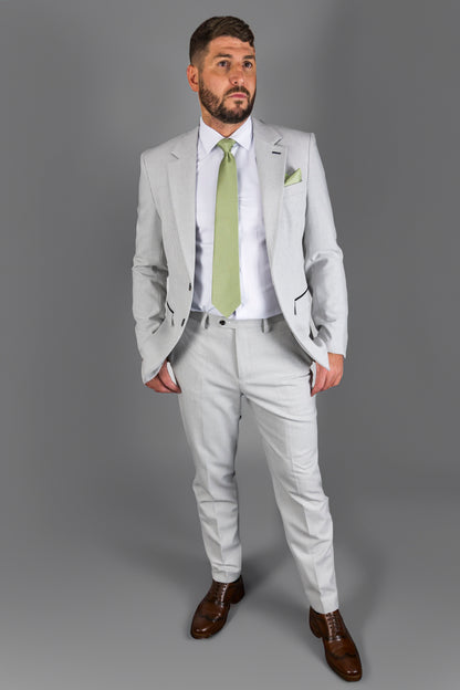 A man wearing the Suitbae Charles stone two-piece suit featuring a classic Prince of Wales check pattern. The outfit includes a tailored jacket and matching trousers paired with a crisp white shirt and a light green tie. Finished with a matching green pocket square and brown formal shoes, creating a sophisticated look perfect for formal events and weddings.