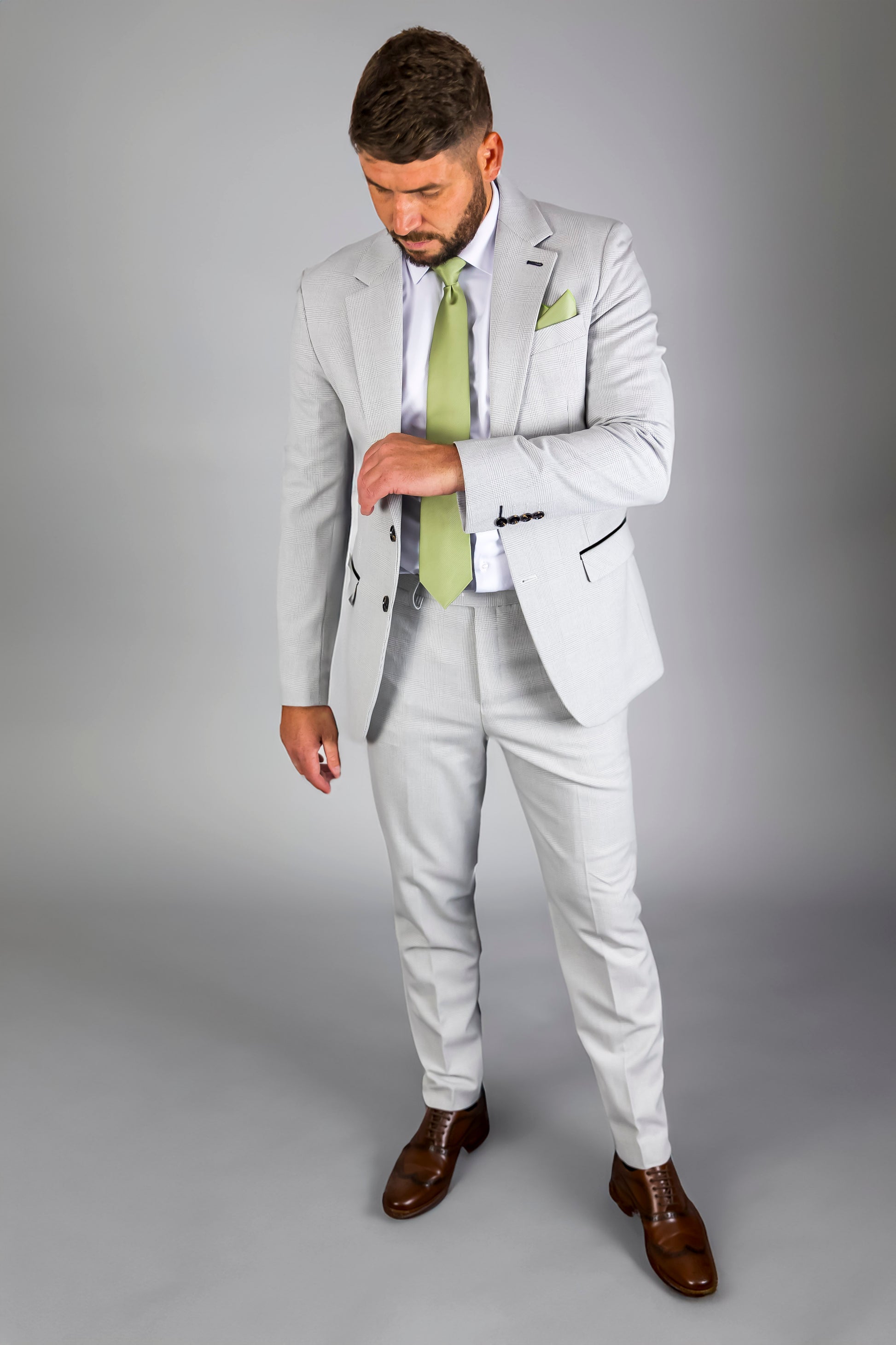 Model wearing a Suitbae Charles stone 2-piece suit with a Prince of Wales check pattern. The suit features a tailored fit, notch lapel, and light green tie paired with a matching pocket square. Styled with brown dress shoes, this outfit offers a refined and modern look for formal occasions. Ideal for big and tall sizes.