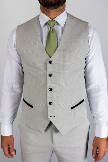 Close-up of a model wearing a Suitbae Charles stone waistcoat paired with a white shirt and a light green tie. The waistcoat features a Prince of Wales check pattern, black contrast piping on the pockets, and a tailored fit, showcasing classic British tailoring with a contemporary twist.