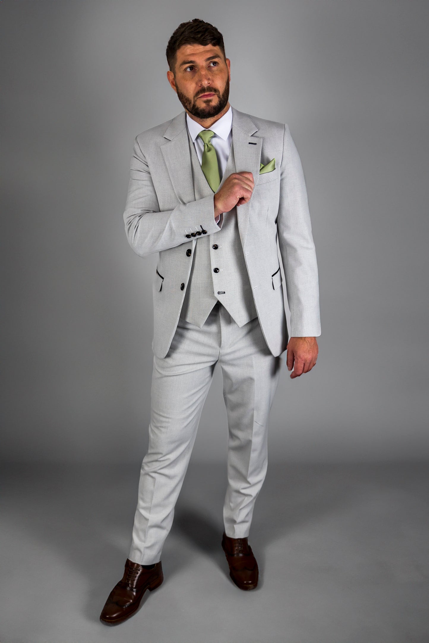 Model dressed in a Suitbae Charles stone three-piece suit, featuring a Prince of Wales check pattern in light grey. Paired with a crisp white shirt, light green tie, and matching pocket square, this tailored look is accented with slim-fit trousers and brown dress shoes. The model adjusts the pocket square, highlighting the refined details and classic fit of the suit.