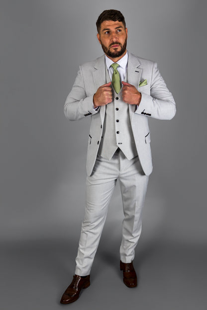 Model wearing the Suitbae Charles stone suit with a light grey Prince of Wales check pattern. The ensemble features a three-piece design, with a matching waistcoat and slim-fit trousers. Paired with a white shirt and a light green tie and pocket square, the look is completed with brown formal shoes. The model adjusts his waistcoat, showcasing the suit’s elegant details.