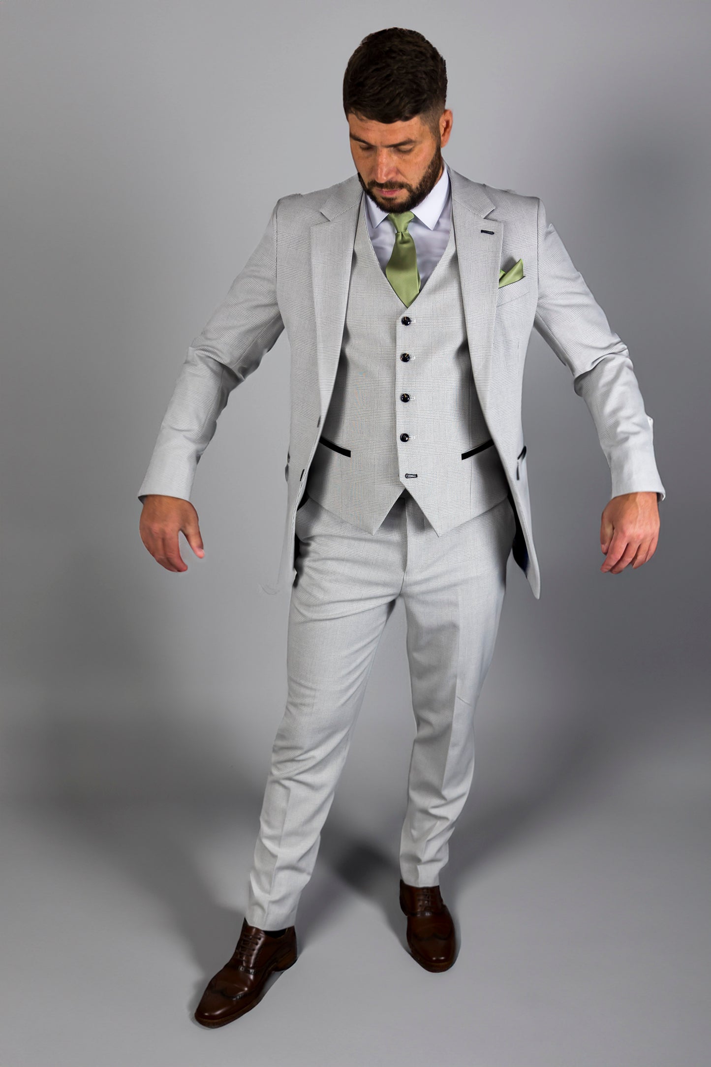 Full-body image of a man wearing the Suitbae Charles stone three-piece suit with a subtle Prince of Wales check pattern. The suit includes a tailored jacket, matching waistcoat, and trousers paired with a crisp white shirt and a light green tie. The ensemble is complemented by brown formal shoes, highlighting a sophisticated and modern style ideal for weddings and formal events.






