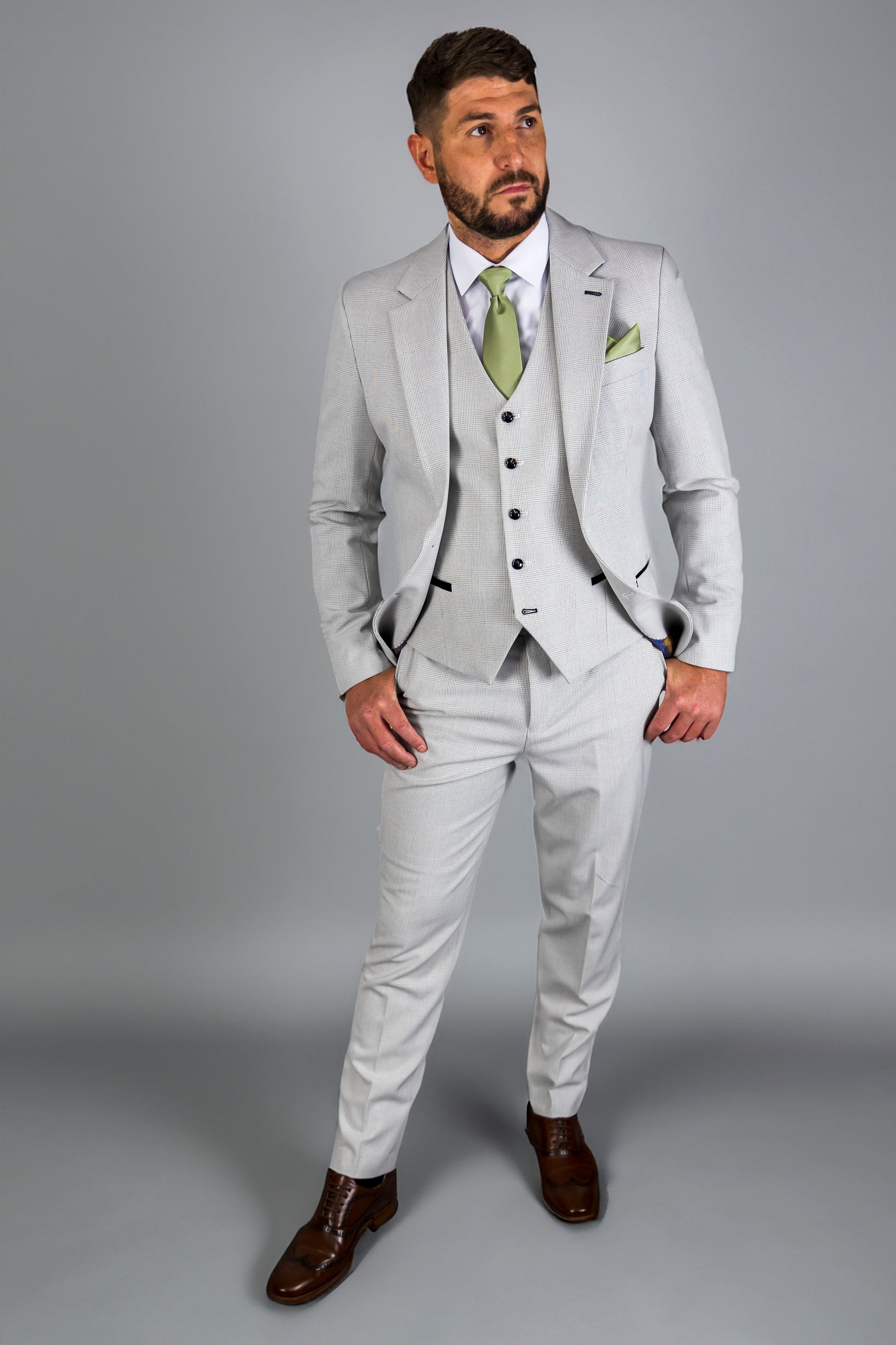 Model wearing a Suitbae Charles stone three-piece suit featuring a Prince of Wales check pattern. The suit is styled with a white shirt, light green tie, and matching pocket square, exuding a modern and sophisticated look. The tailored fit and slim-cut trousers are complemented by polished brown dress shoes, creating an elegant ensemble suitable for formal occasions.