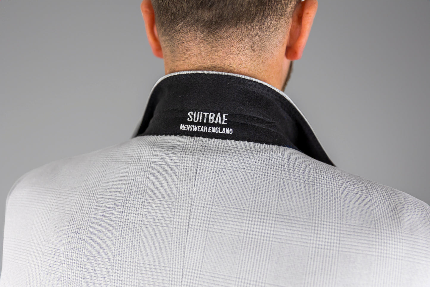 Close-up of the back collar of a Suitbae Charles stone suit, showcasing the "Suitbae Menswear England" embroidered branding on a black velvet collar lining, set against the light grey Prince of Wales check fabric.