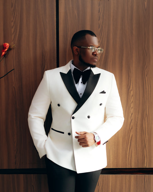 A man wearing a white double-breasted tuxedo jacket with black satin lapels, a black bow tie, and a white shirt, standing against a dark wood-paneled wall.