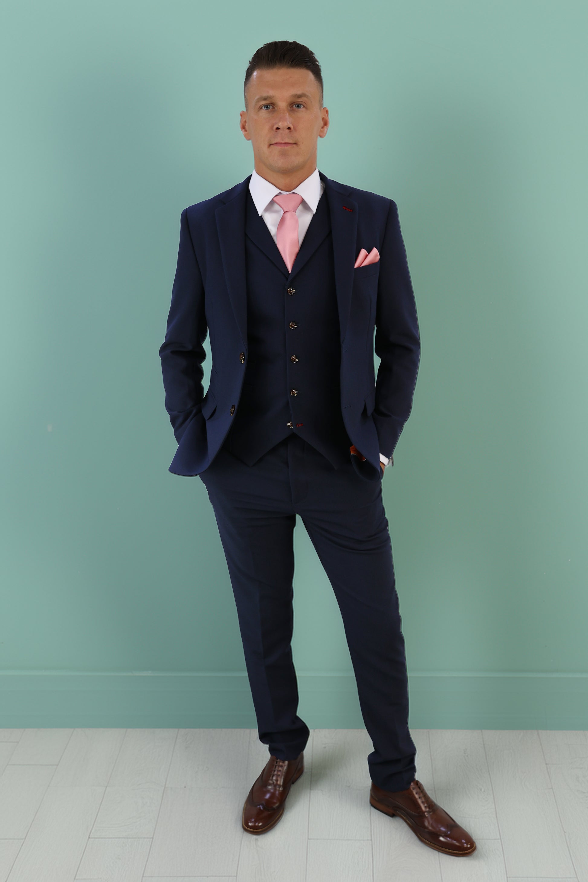 Model wearing a tailored navy three-piece suit with a pink tie and pocket square, styled with brown leather shoes, showcasing Suitbae's expert tailoring for XL, Plus Size, and Big & Tall men's sizes. Shop suits, shirts, ties, and accessories at Suitbae.com.