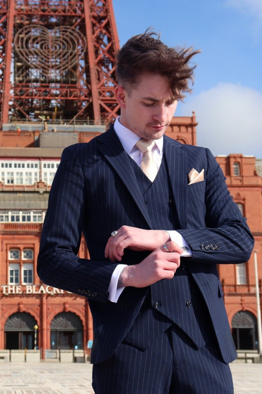 Man adjusting his cuff in front of Blackpool Tower wearing a navy pinstripe three-piece suit over a white shirt, styled with a champagne tie and matching pocket square — part of the SUITBAE collection.