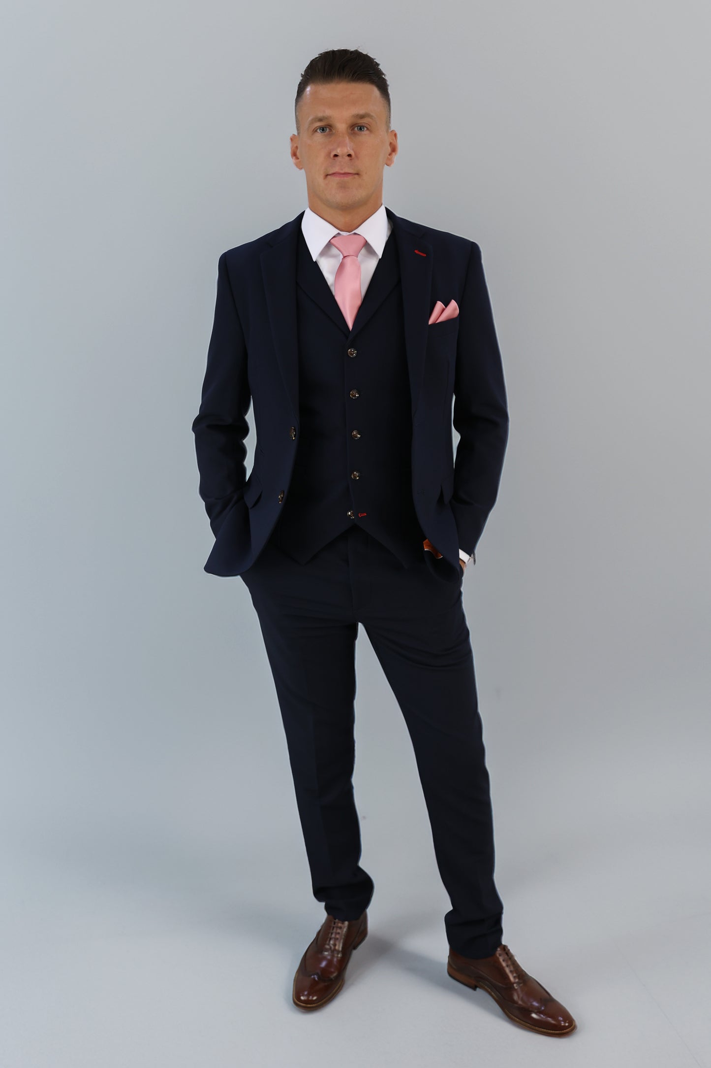 Model in a navy three-piece suit with a pink tie and pocket square, styled with brown leather shoes. Expertly tailored for XL, Plus Size, and Big & Tall men's sizes. Shop premium menswear at Suitbae.com