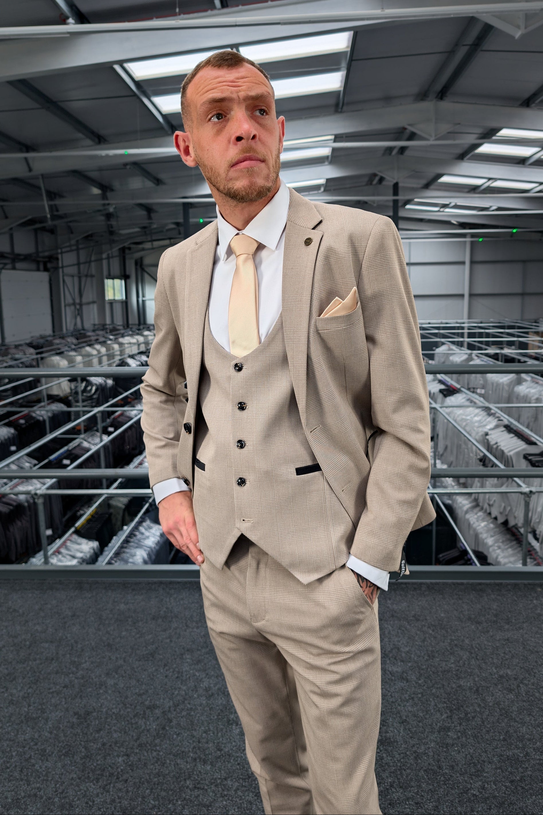 A man modeling a Suitbae beige 3 piece suit with a subtle checkered pattern, styled with a white shirt and cream tie in a clothing warehouse.